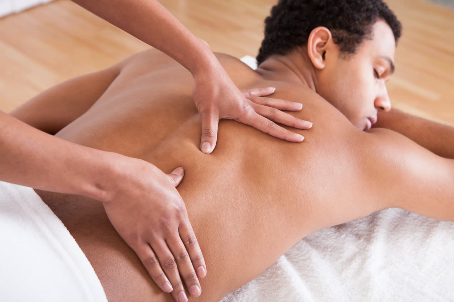 A man lies on a massage table with his eyes closed and a towel covering him from the waist down while two hands massage his back.